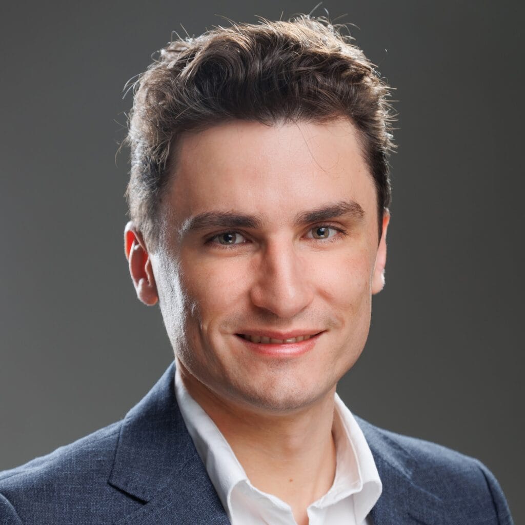 Um homem jovem de pele clara, cabelo castanho curto e penteado para cima, vestindo um blazer cinza e camisa branca. Ele está sorrindo levemente e olhando para a câmera, com um fundo cinza neutro.